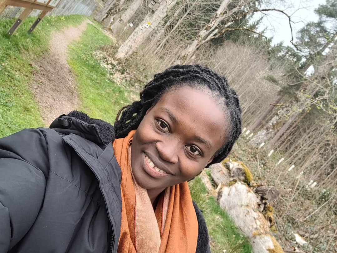 Eine lächelnde Frau macht ein Selfie auf einem Waldweg und trägt eine schwarze Jacke sowie einen orangefarbenen Schal.