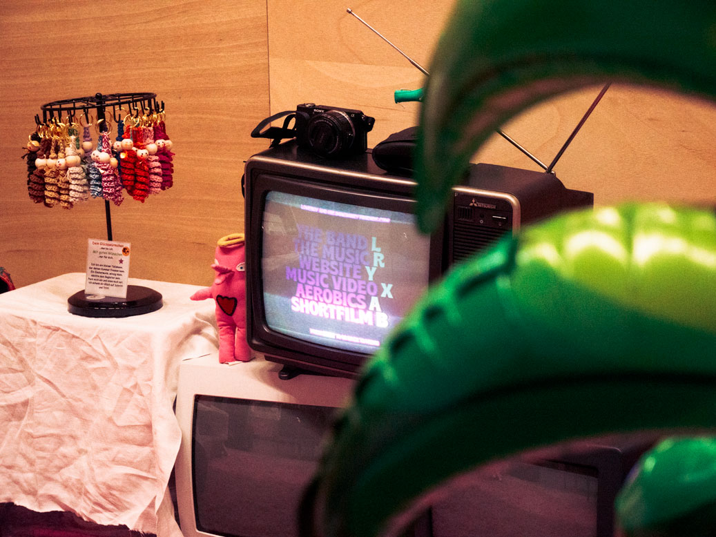 Nicolaj Ésteban's 'Loveboy and His Imaginary Friends' booth at Herofest 2024 in Bern. A vintage television displays the band's name and links to their website, music, videos, and a short film. Merchandise, including a plush toy and keychains, is also visible.