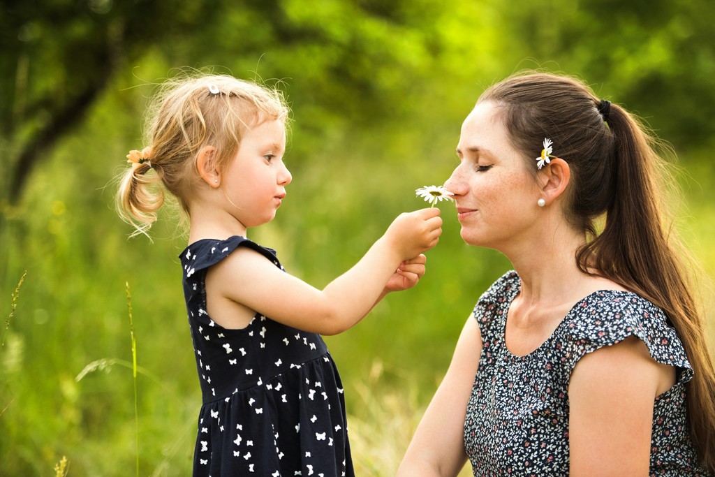 A young girl offers a daisy to her smiling mother, both dressed in matching floral outfits, sharing a tender moment in a lush green field.