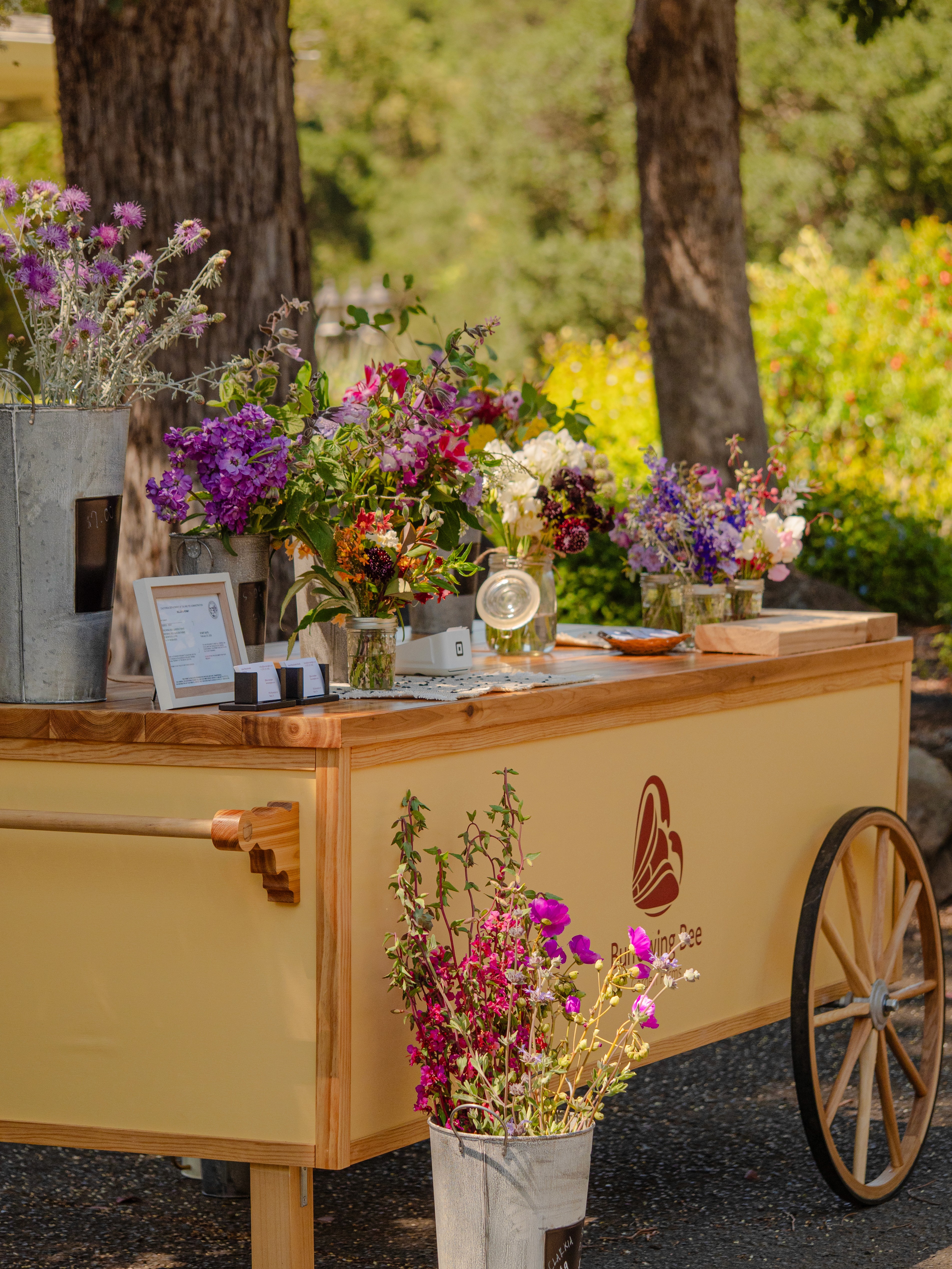 A photo of the Burrowing Bee Garden Farms flower cart stocked with a wide variety of flowers. The cart is yellow and natural wood with small wagon wheels.