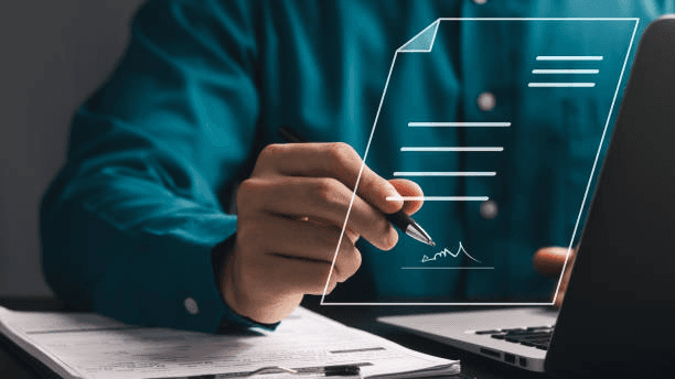 Een persoon in een blauwgroen shirt bekijkt documenten terwijl hij een laptop gebruikt en een transparant scherm met gegevens vasthoudt.