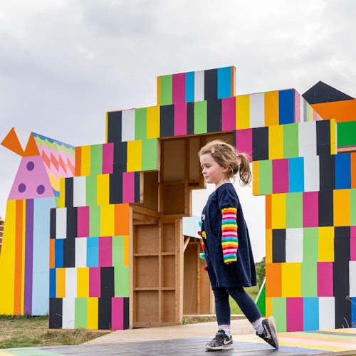 Ein Kind läuft draußen vor einer großen, farbenfrohen Blockstruktur unter einem bewölkten Himmel.