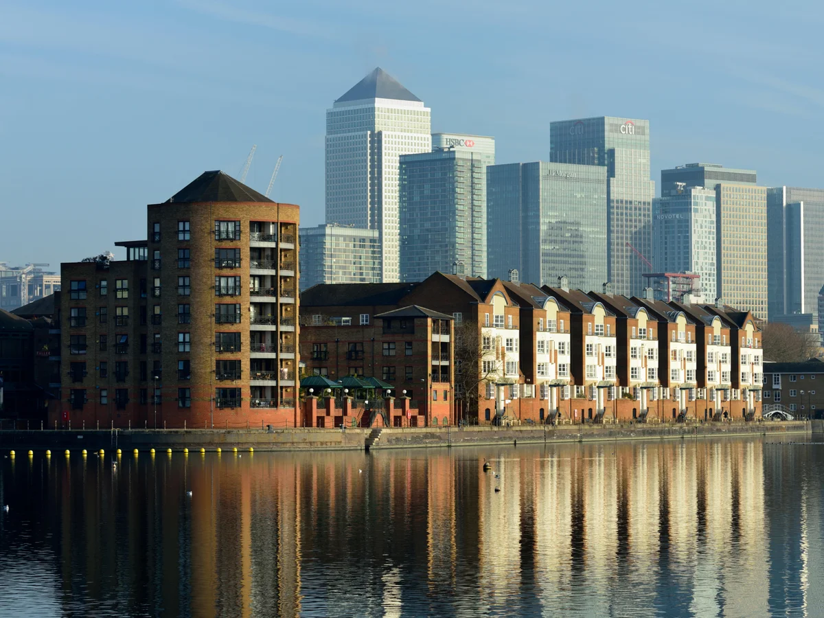 A smiling man with curly hair is sitting at a desk, looking at the camera, with a laptop and a lamp in the background.
