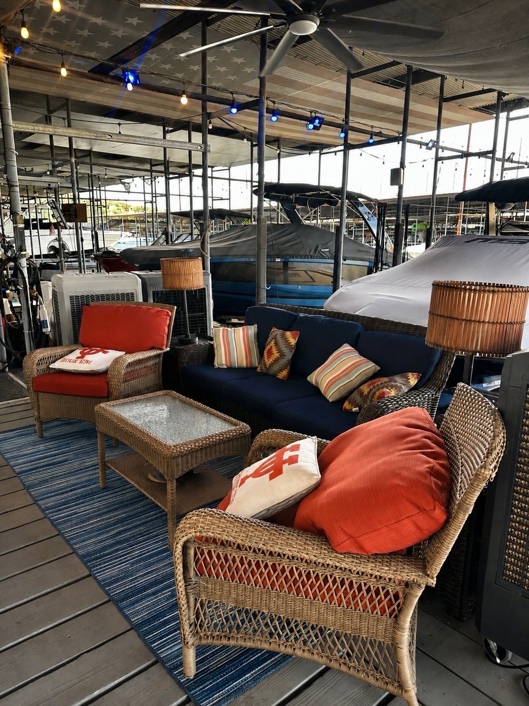 A cozy seating area on a dock features a blue sofa with patterned pillows, wicker chairs with red cushions, a wicker coffee table, and soft lighting, set against a backdrop of moored boats.