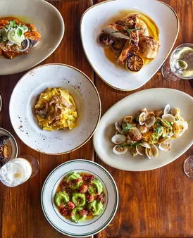Overhead view of four elegant Italian dishes on a wooden table, including pasta, seafood, and a green appetizer, accompanied by a drink.