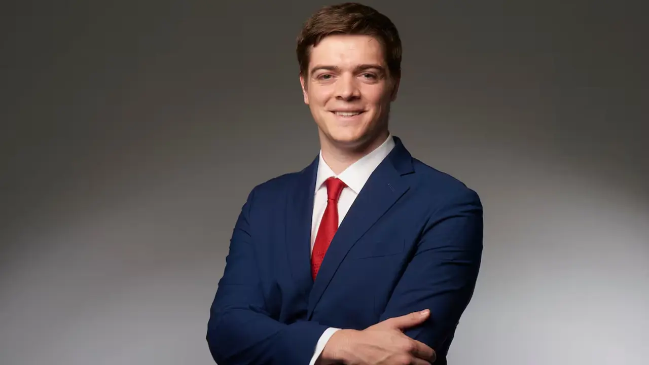 Retrato de estudio de un hombre joven de traje azul y corbata roja, con los brazos cruzados sobre fondo gris.