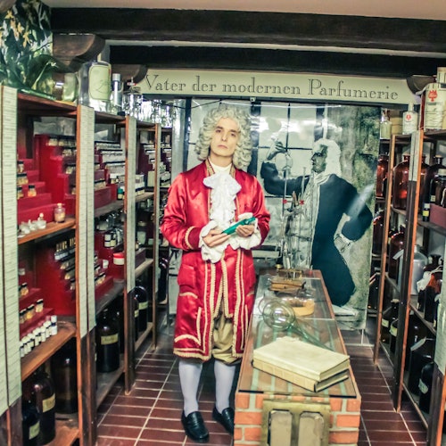 A wax figure in period attire stands in a historical perfume shop, surrounded by shelves of bottles and books.