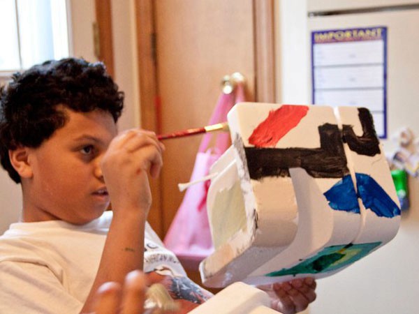 A young boy with dark curly hair, wearing a white t-shirt, is painting a white rectangular block with black, red, and blue paint. He holds a red-handled paintbrush in his right hand and applies the paint to the block. The block rests on a white surface. In the background, a pink towel hangs on a hook, and a white wall displays a blue sign with the word “IMPORTANT” visible.