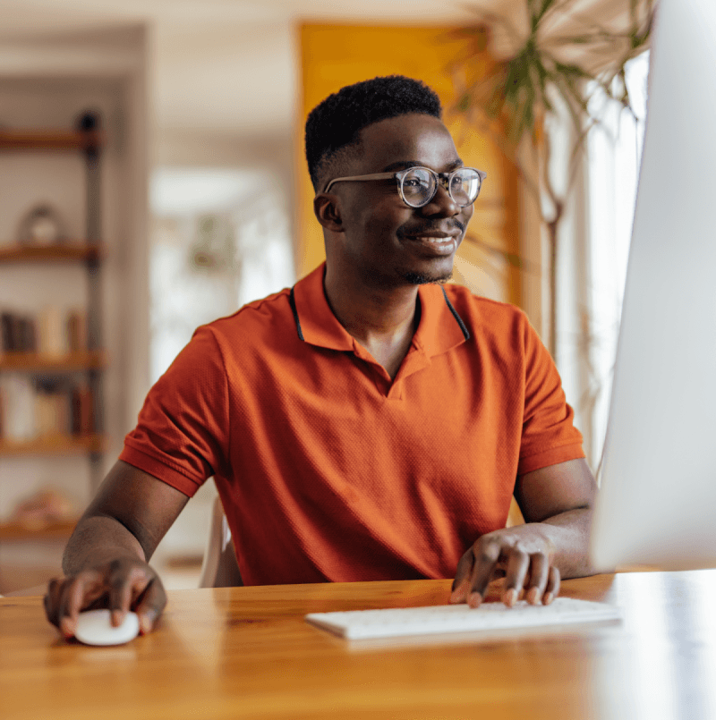 A man in an orange shirt sitting at a desk with a computer and mouse.