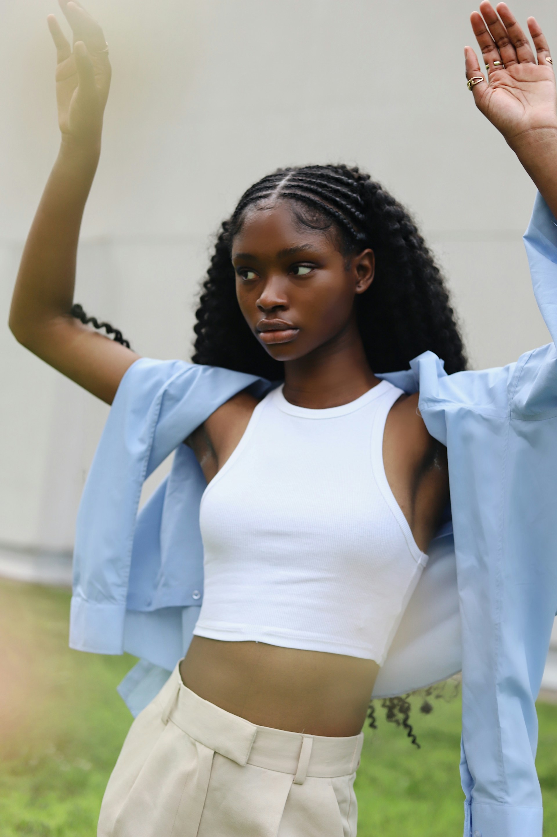 A young Black woman posing with both hands raised, looking to the side. She wears a white ribbed crop top and light-colored trousers with a blue shirt draped loosely over her shoulders against a blurred outdoor background.