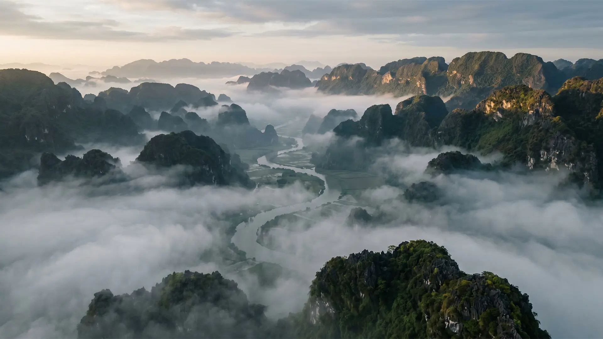 Aerial view of mist-covered limestone karst formations and winding river valleys in Ninh Binh, Vietnam, at sunrise.