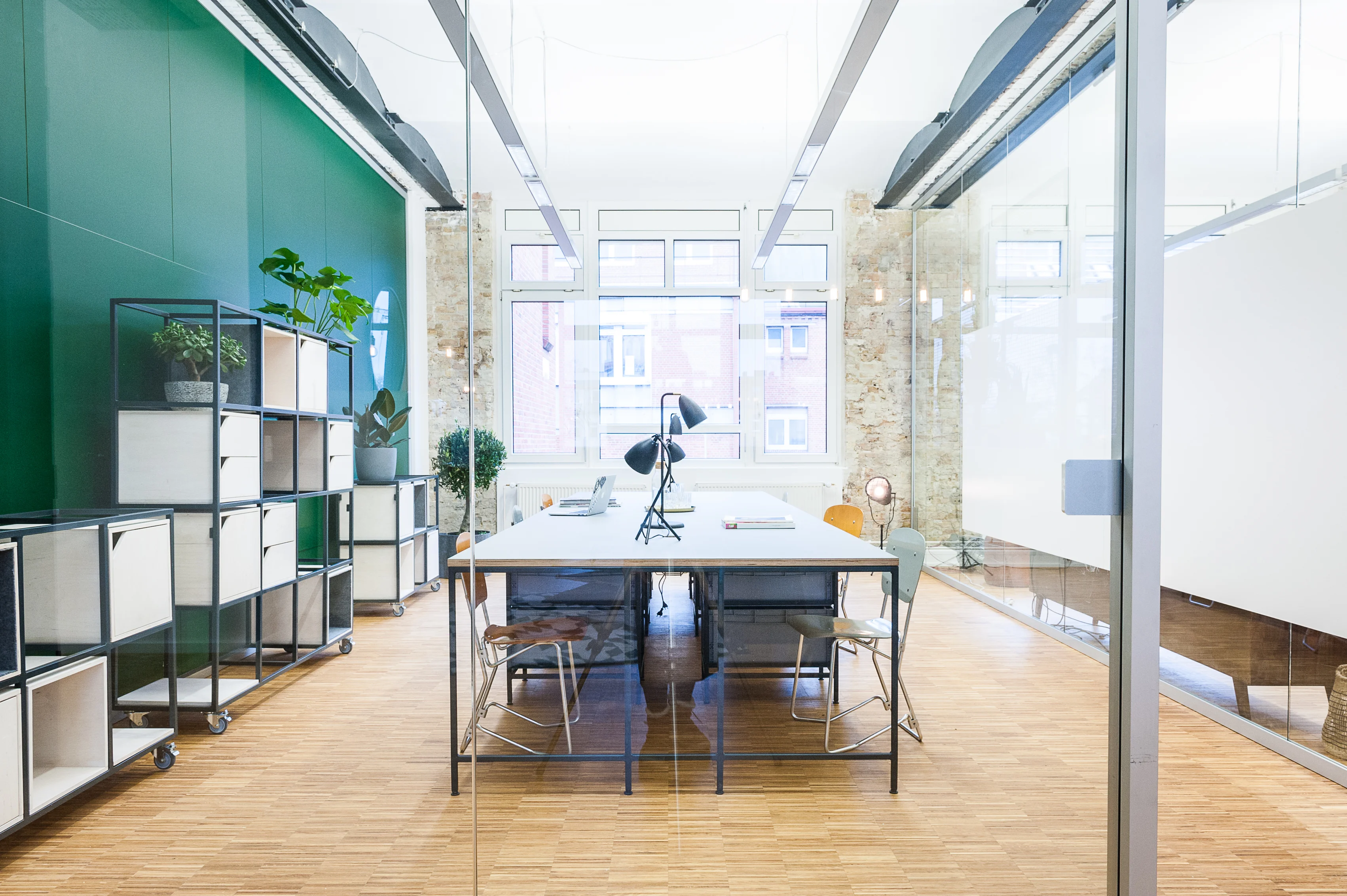 Office room at INTERMATE x TRUEMATES with a glossy green wall, modular shelving on castors, a shared white desk with black task lamps and exposed brick walls behind glass partitions.