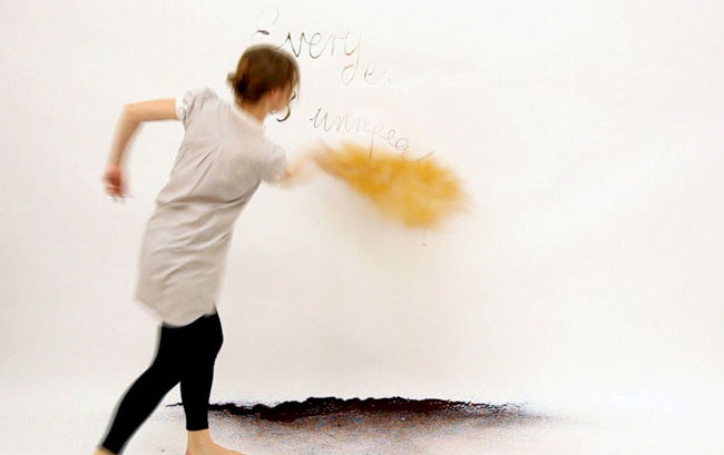 A person with short brown hair, wearing a light gray short-sleeved shirt and black leggings, is captured mid-motion against a white wall. Their right arm is extended forward, and their left arm is bent behind them. A yellow, cloud-like shape is visible near their right hand. Above them, the words “Every day…unprepare” are written in black marker on the wall. The ground in front of the person appears dark and uneven.