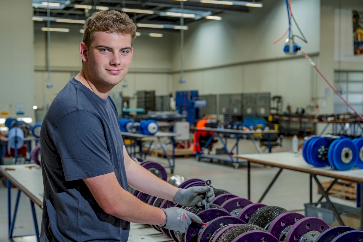 Ein junger Mann lächelt, während er ein Teil in einer Fabrik hält. Teile auf einem Tisch, Werkzeuge, Maschinen und andere Arbeiter.