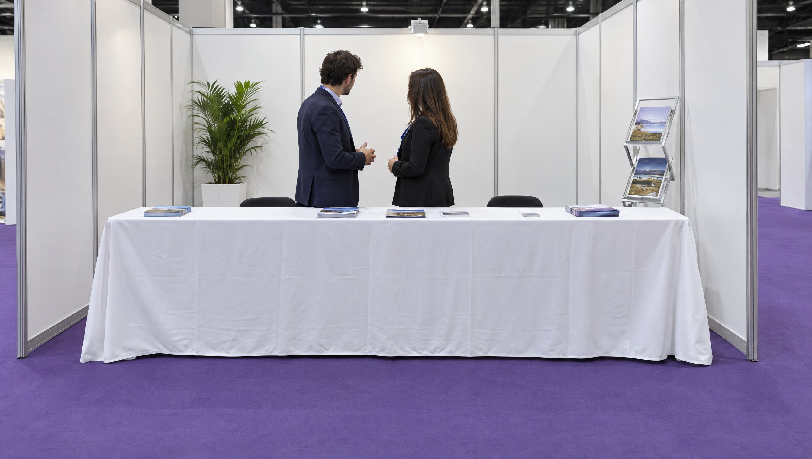 Twee standhouders staan met hun rug naar het gangpad achter een lange tafel met wit kleed, die de voorkant van de beursstand afsluit
