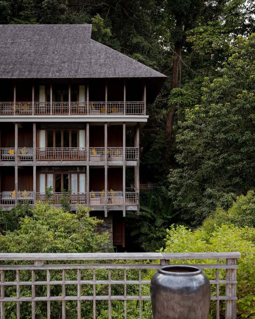 Exterior view of a multi-story wooden resort building surrounded by lush green rainforest, showcasing balconies with yellow chairs.