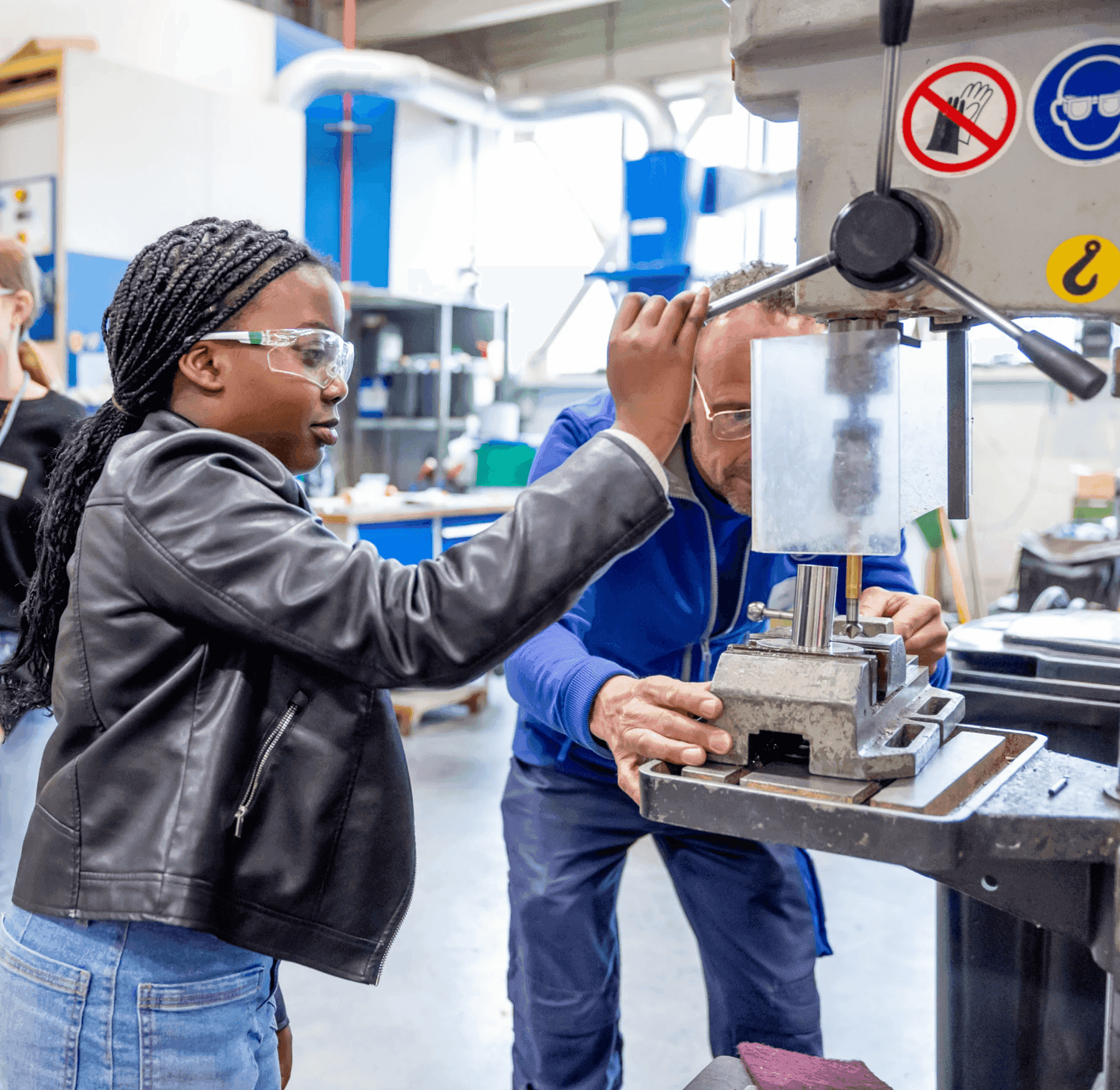 Young female student in safety glasses operating a drill press, guided by a male instructor at a vocational training workshop
