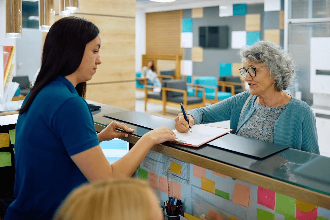 An elderly woman with gray hair signs a document at a modern reception desk, attended by a staff member in a blue uniform, with colorful sticky notes lining the front of the desk in a contemporary office environment.