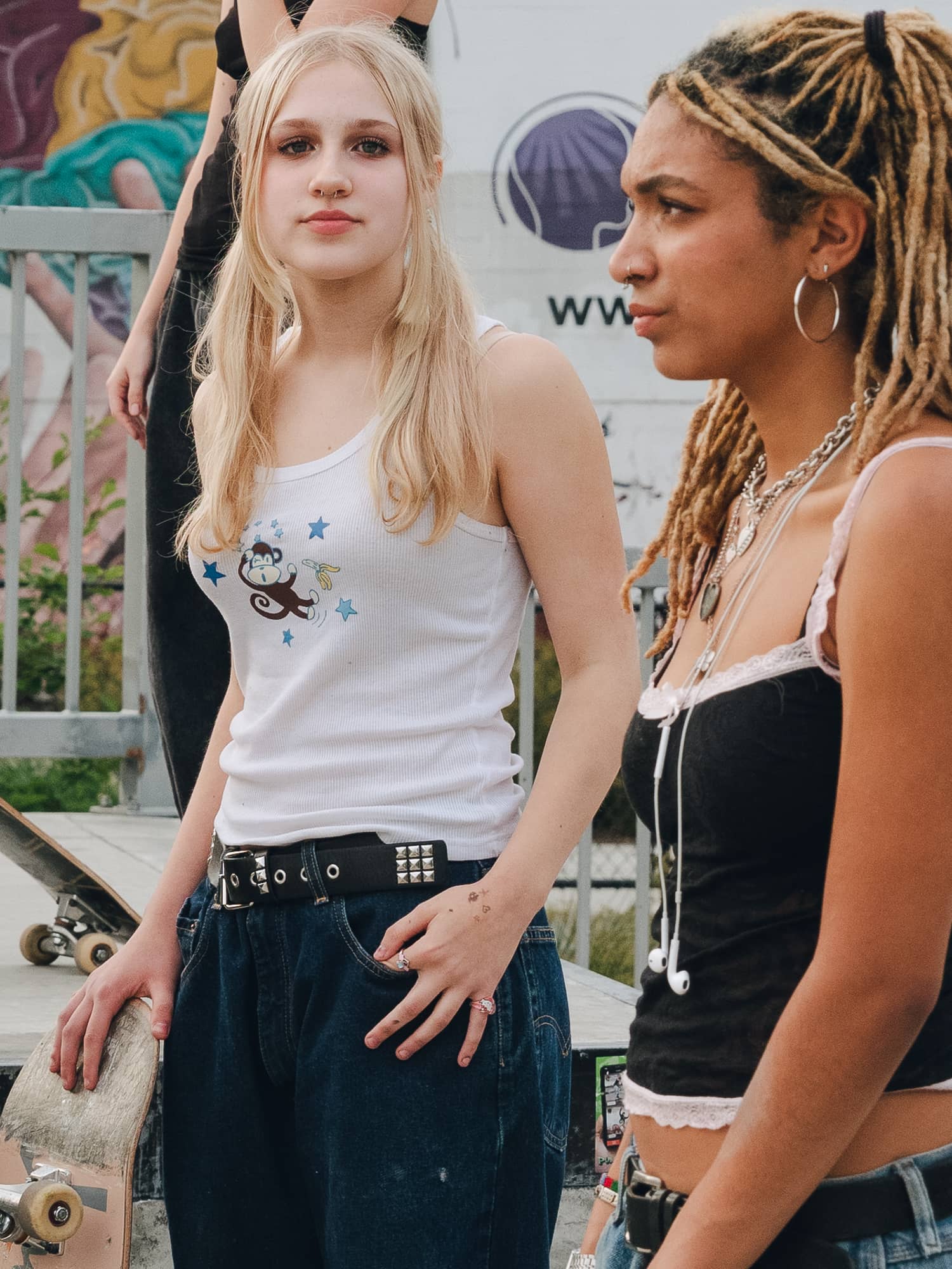 Two young women at a skate park, one holding a skateboard. The scene is lively, with vibrant graffiti in the background. They appear relaxed and confident.