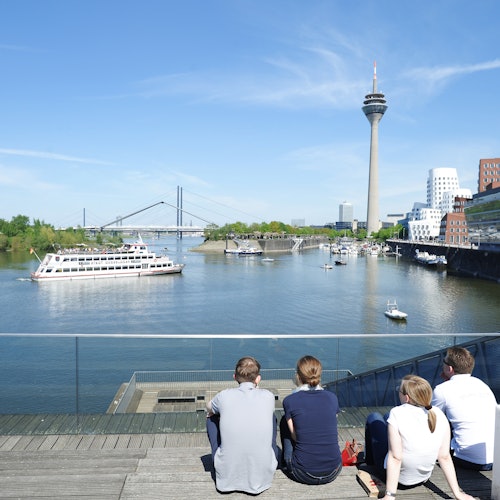 Vier Personen sitzen auf Holzstufen am Hafen, im Hintergrund sind Boote, eine Brücke und ein hoher Aussichtsturm zu sehen.