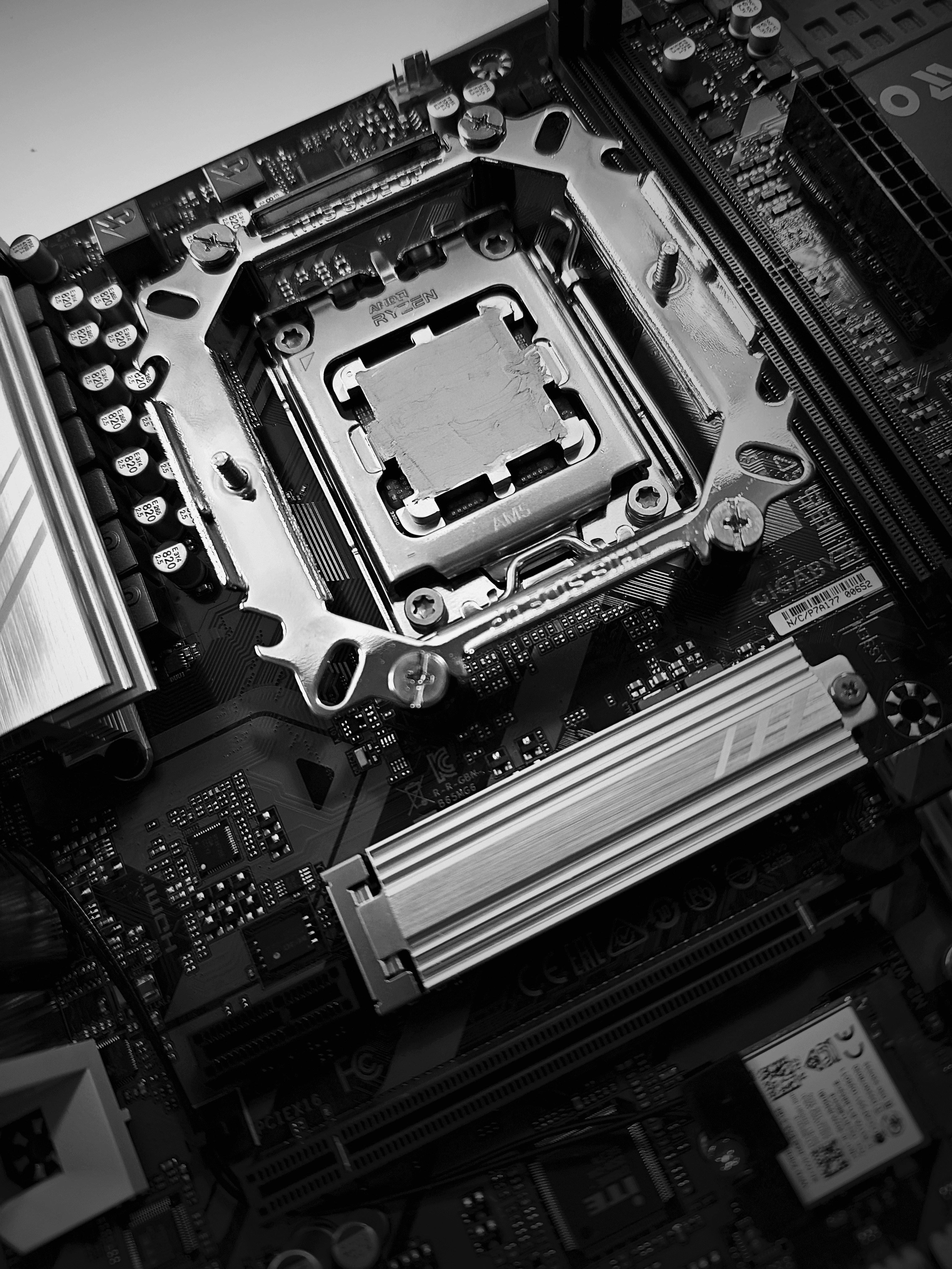 Czarno-białe zdjęcie z bliska przedstawiające procesor AMD Ryzen osadzony w gnieździe AM5 na płycie głównej Gigabyte. Na środku procesora widoczna jest świeżo nałożona warstwa pasty termoprzewodzącej; w tle widoczny radiator sekcji zasilania oraz sloty pamięci RAM.