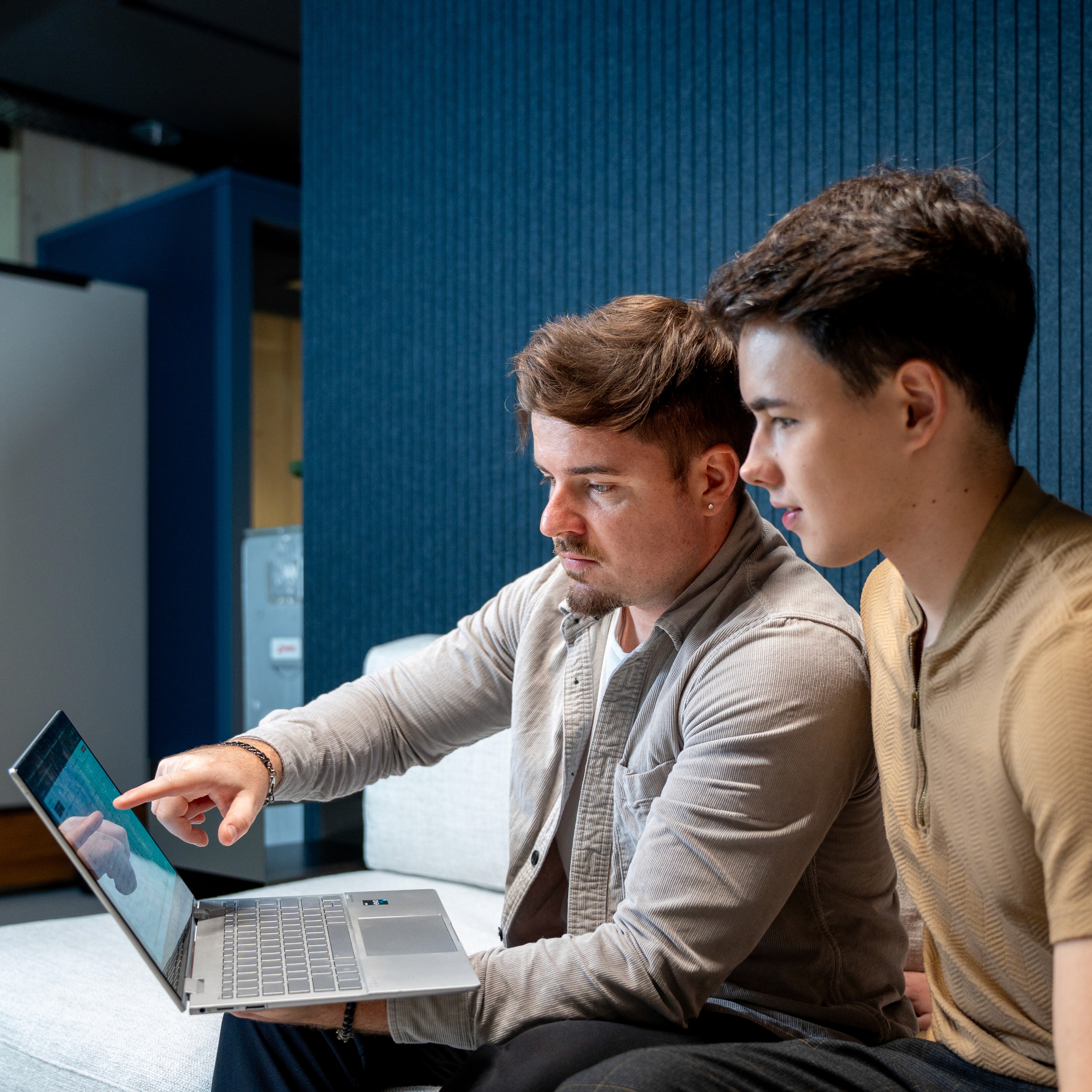 A young person with curly hair smiles at the camera while sitting in front of a laptop against a dark background.