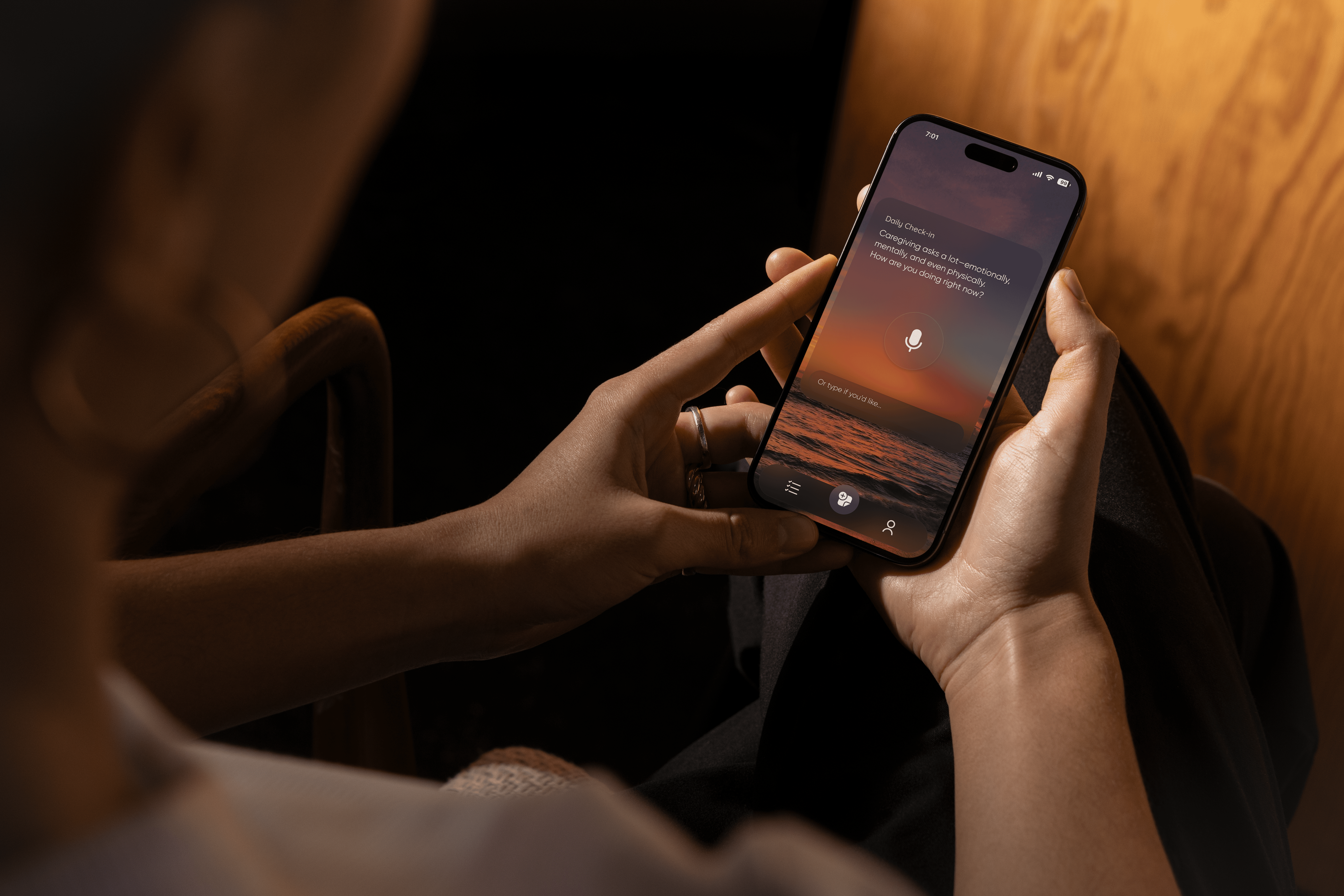 Mobile app screen showing a daily emotional check-in prompt that reads, “What’s something your Mom did today that made you smile?” The app features a microphone button and a sunset background, with the phone held in a hand against a softly lit indoor setting.