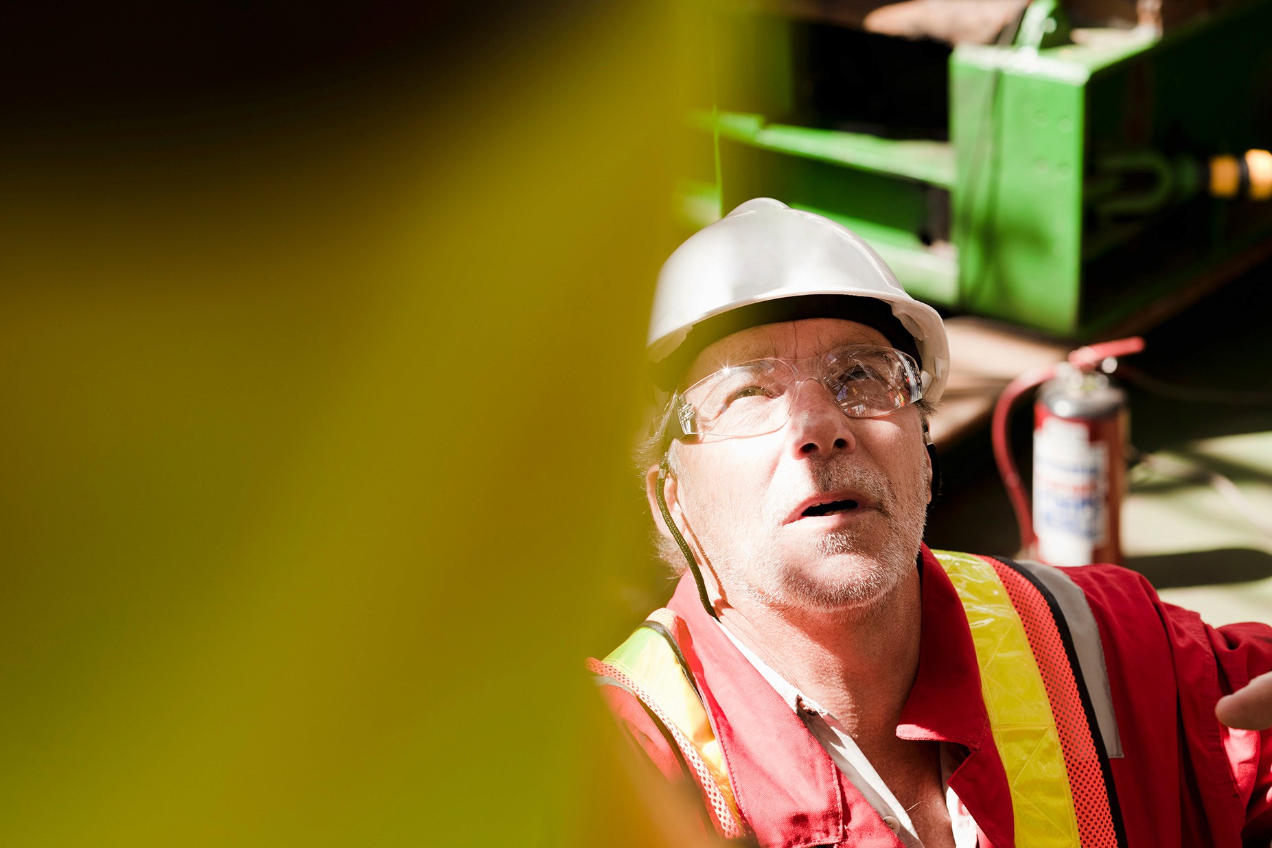 A man wearing safety glasses, a white helmet, and red overalls looks up in a factory environment.