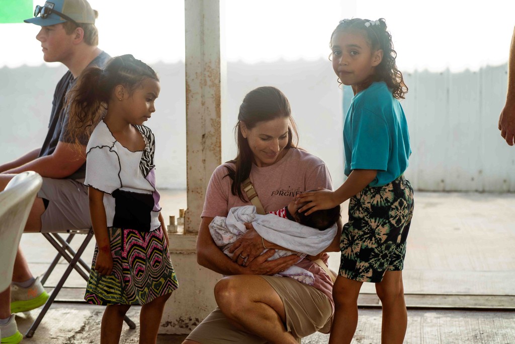 A group of children seated on the floor in a bright, spacious indoor area, with adults supervising.