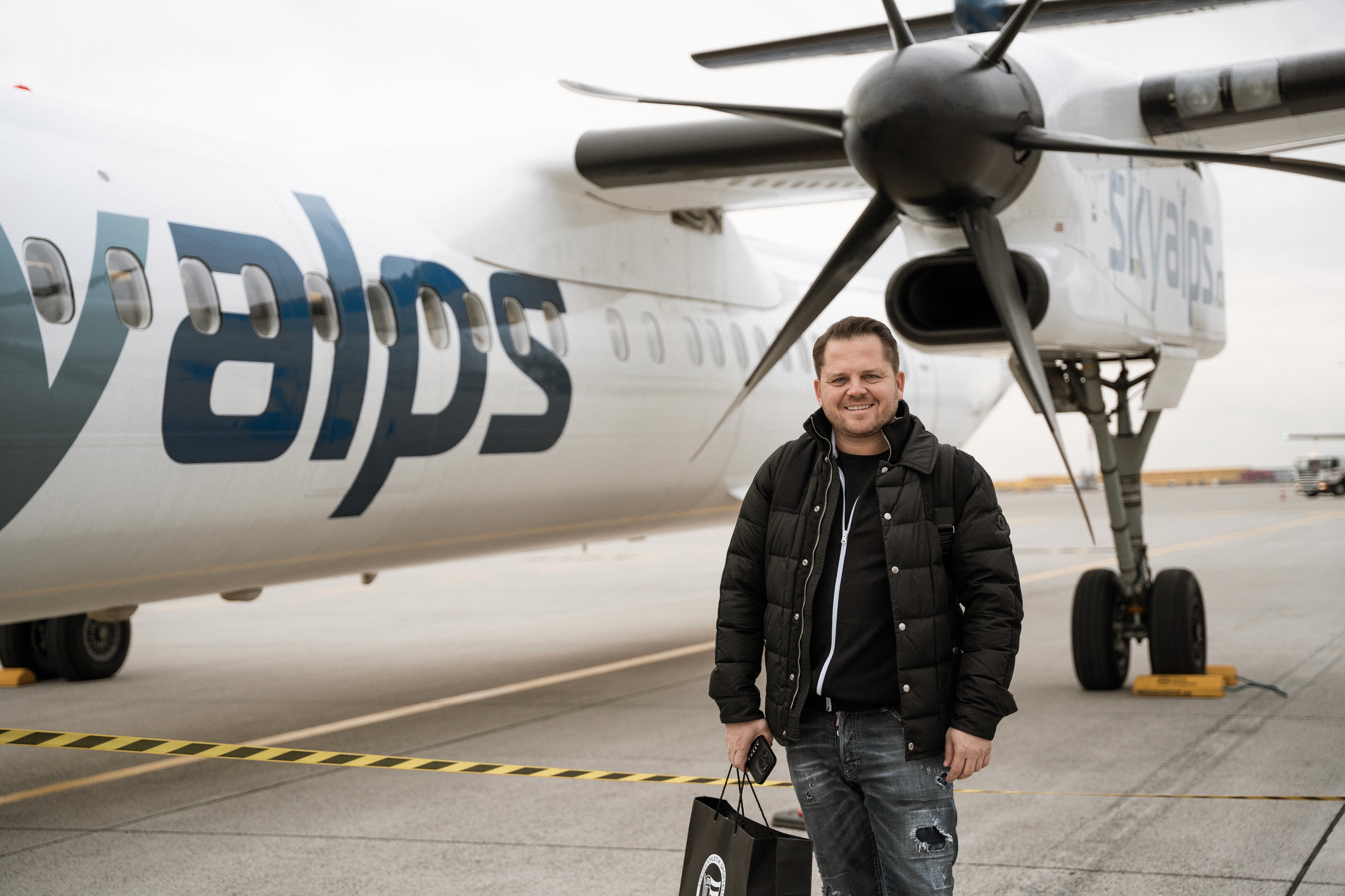 Ein Mann posiert vor dem Flugzeug von Sky Alps mit einem Gepäckstück. Er lächelt in die Kamera.