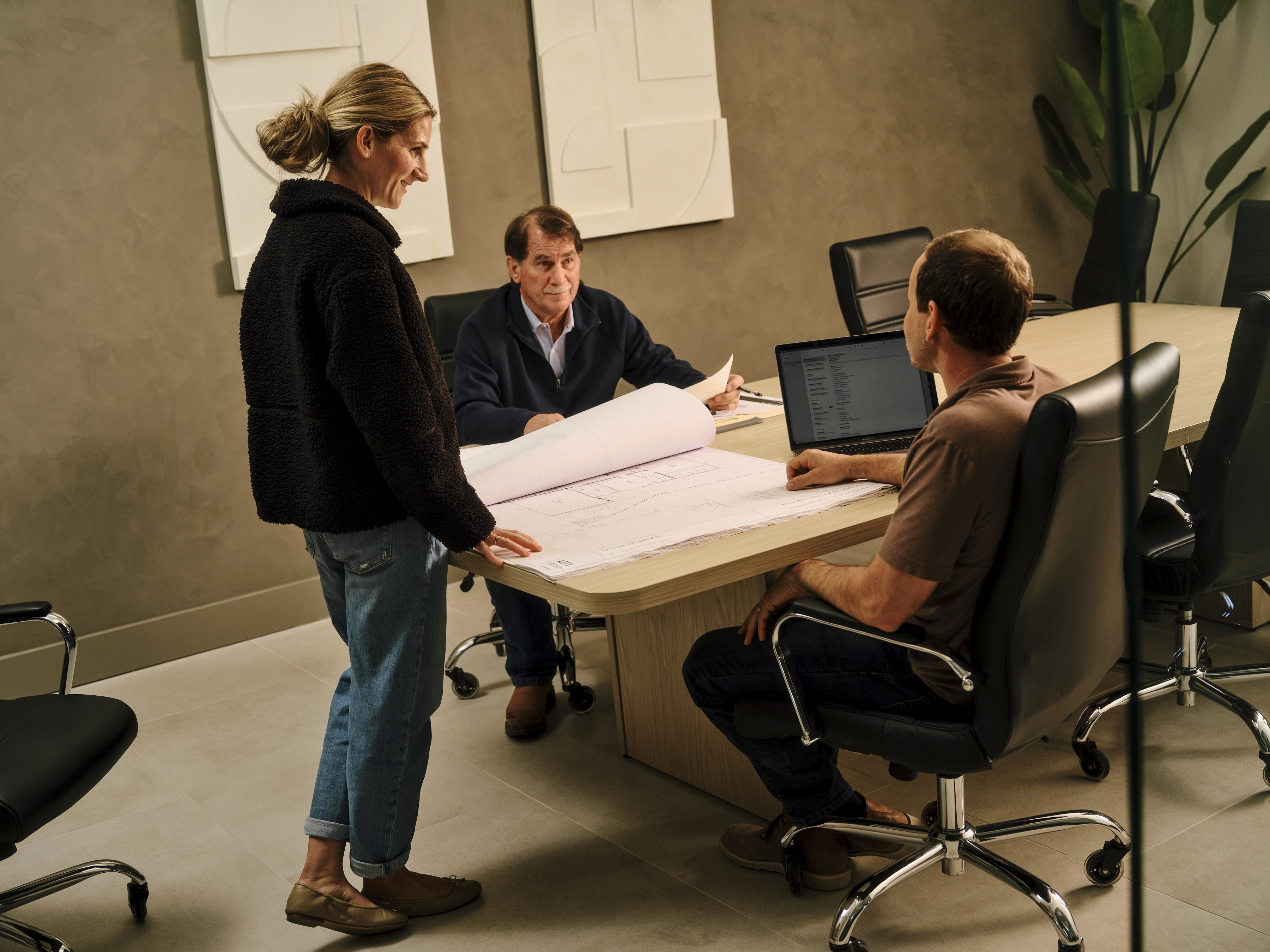 Three professionals in a modern conference room review architectural blueprints spread across a table next to an open laptop