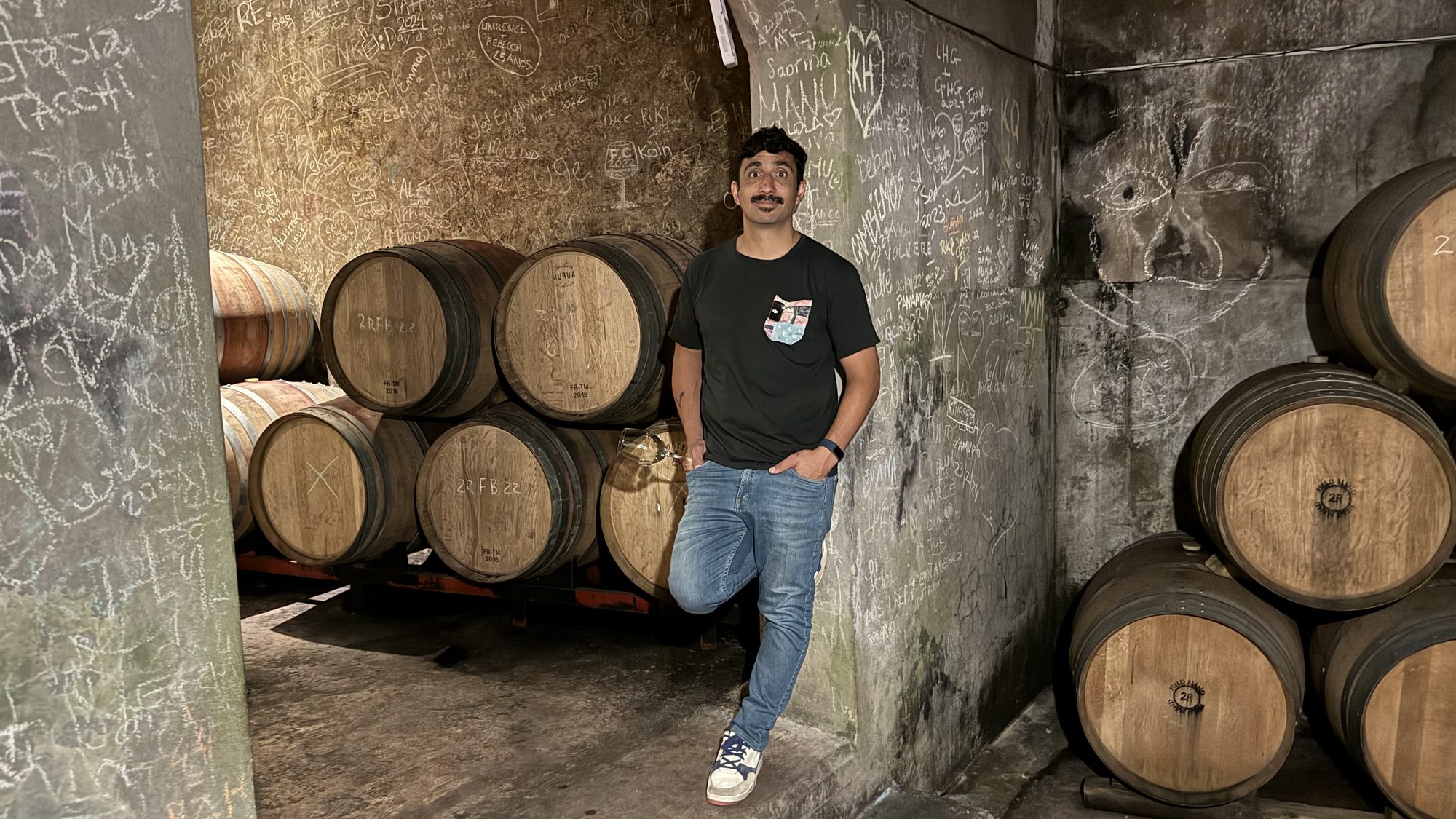 Winemaker Ramiro Maures standing among oak wine barrels in a compact cellar with signatures and drawings covering the concrete walls.