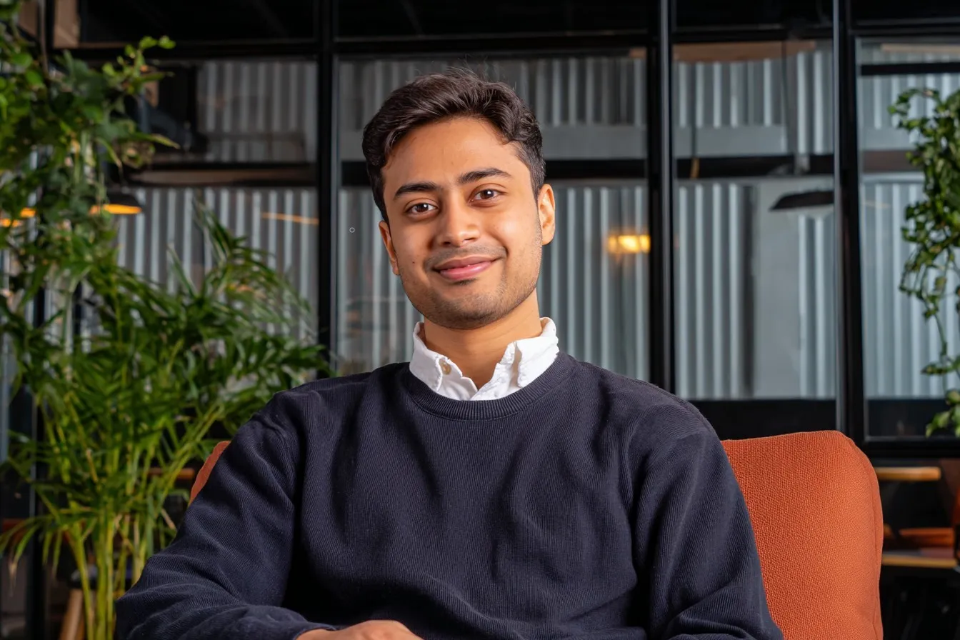 Man in a navy sweater sits in a cafe with plants and glass panels behind.