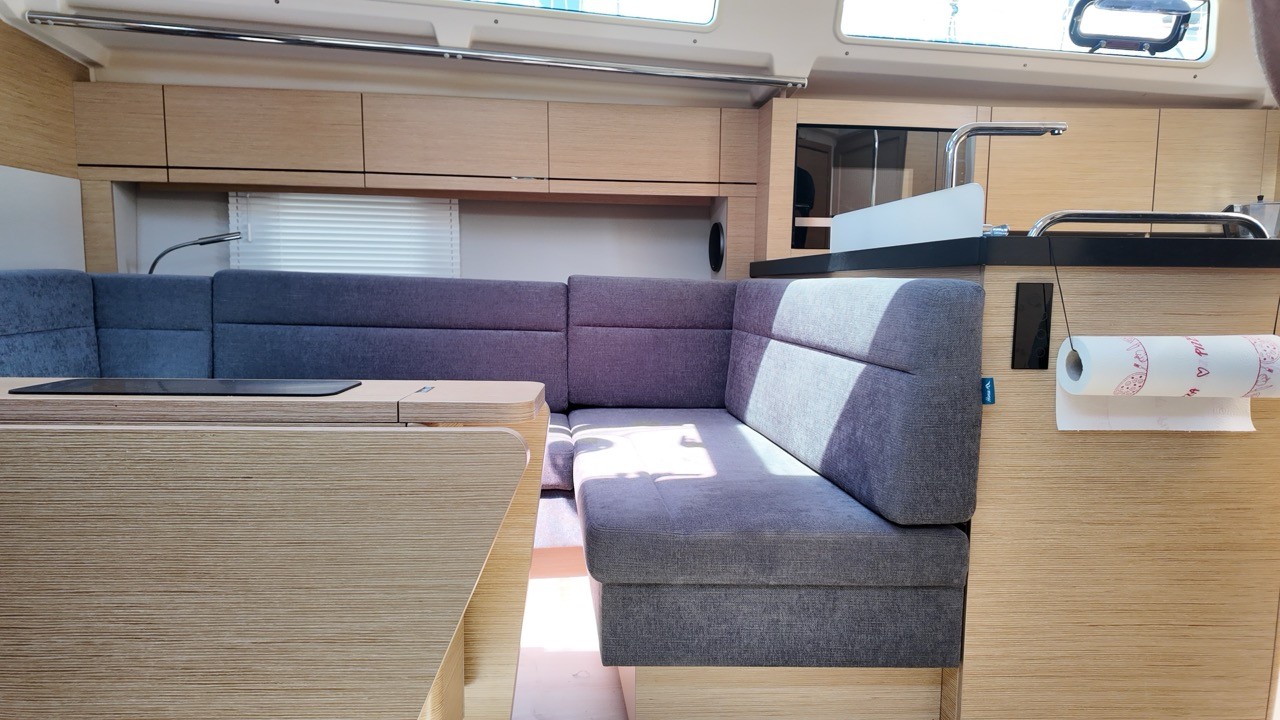 Interior view of the Hanse 388's saloon, showing an L-shaped gray upholstered settee, light oak cabinetry, a fold-out table, and a galley counter with a chrome faucet and paper towel holder.