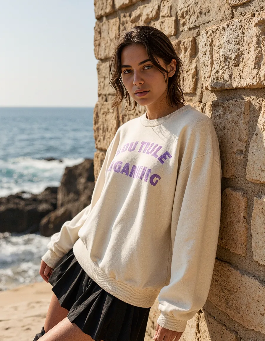 Fashion portrait against stone wall by the sea, featuring cream sweatshirt with purple text and black skirt with ocean backdrop