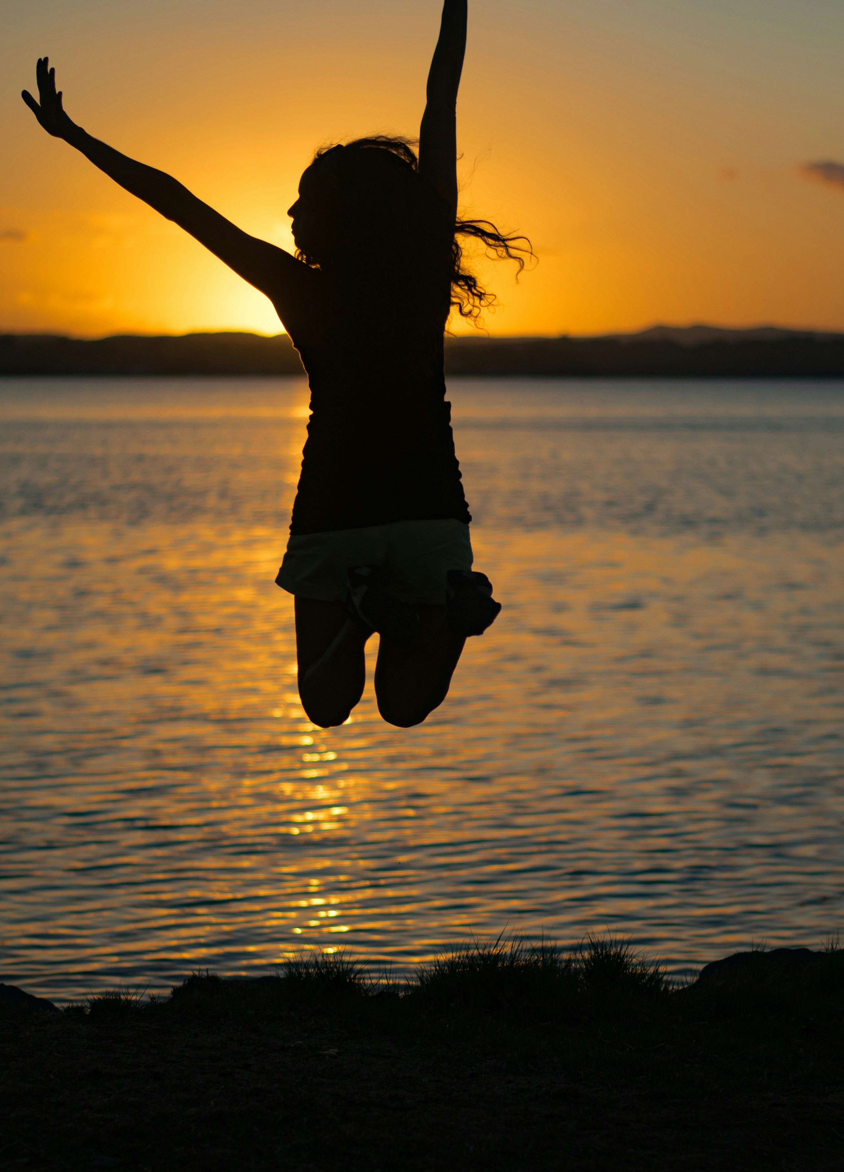 A silhouette of a person jumping joyfully against a stunning sunset over a calm lake, creating a vibrant reflection on the water's surface.