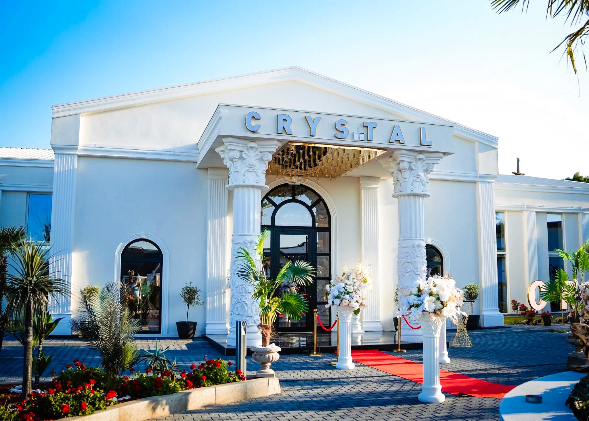 A modern hallway features vibrant teal and orange geometric and leaf patterns, with glossy tiled floors, a large rectangular light fixture, and a small shelf holding a potted plant, all leading to a large, frosted window at the end.