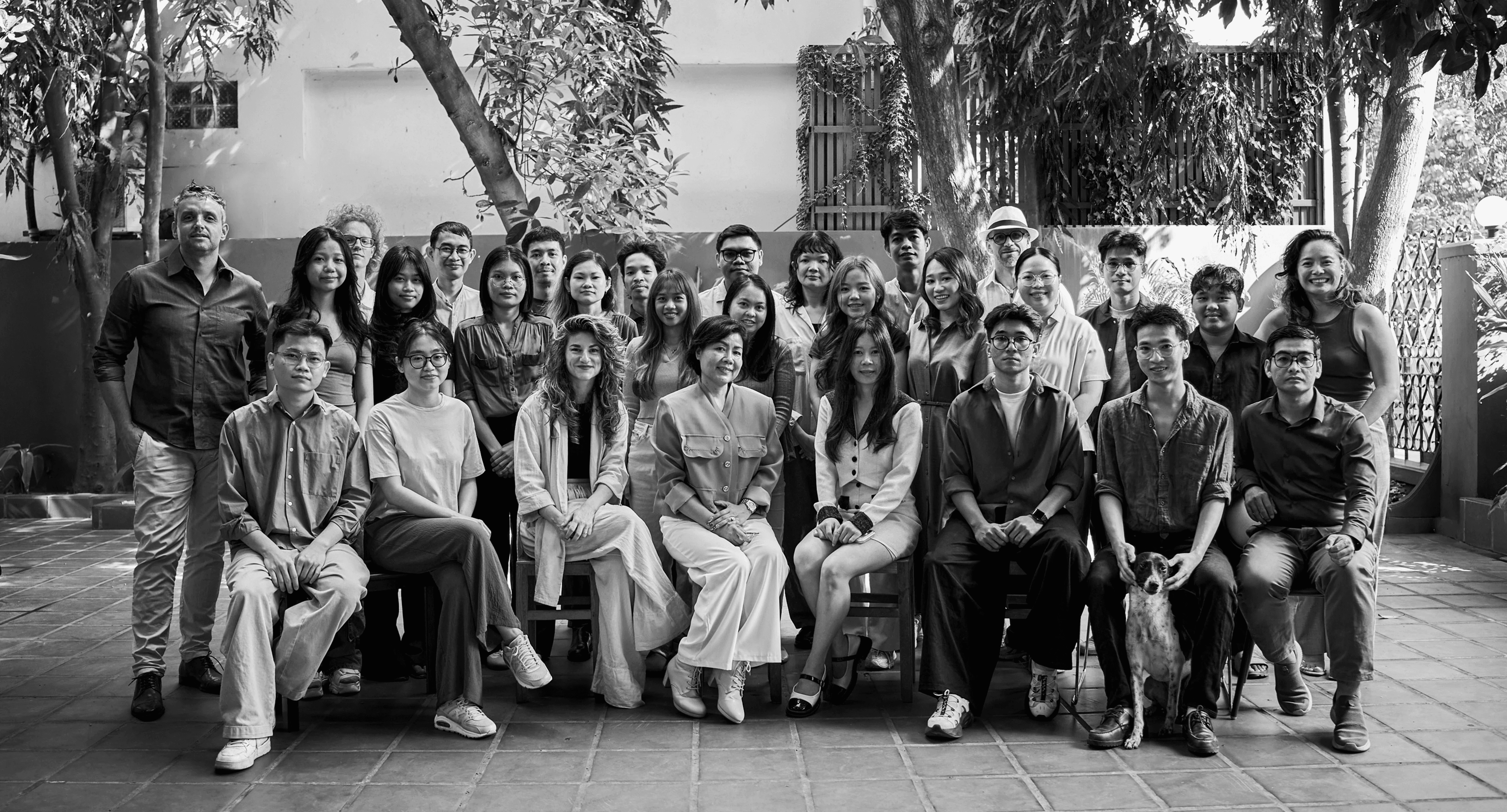 A black and white group photo of The Room Architecture and Design team posing outdoors in a courtyard with trees. The staff is dressed in professional business attire.
