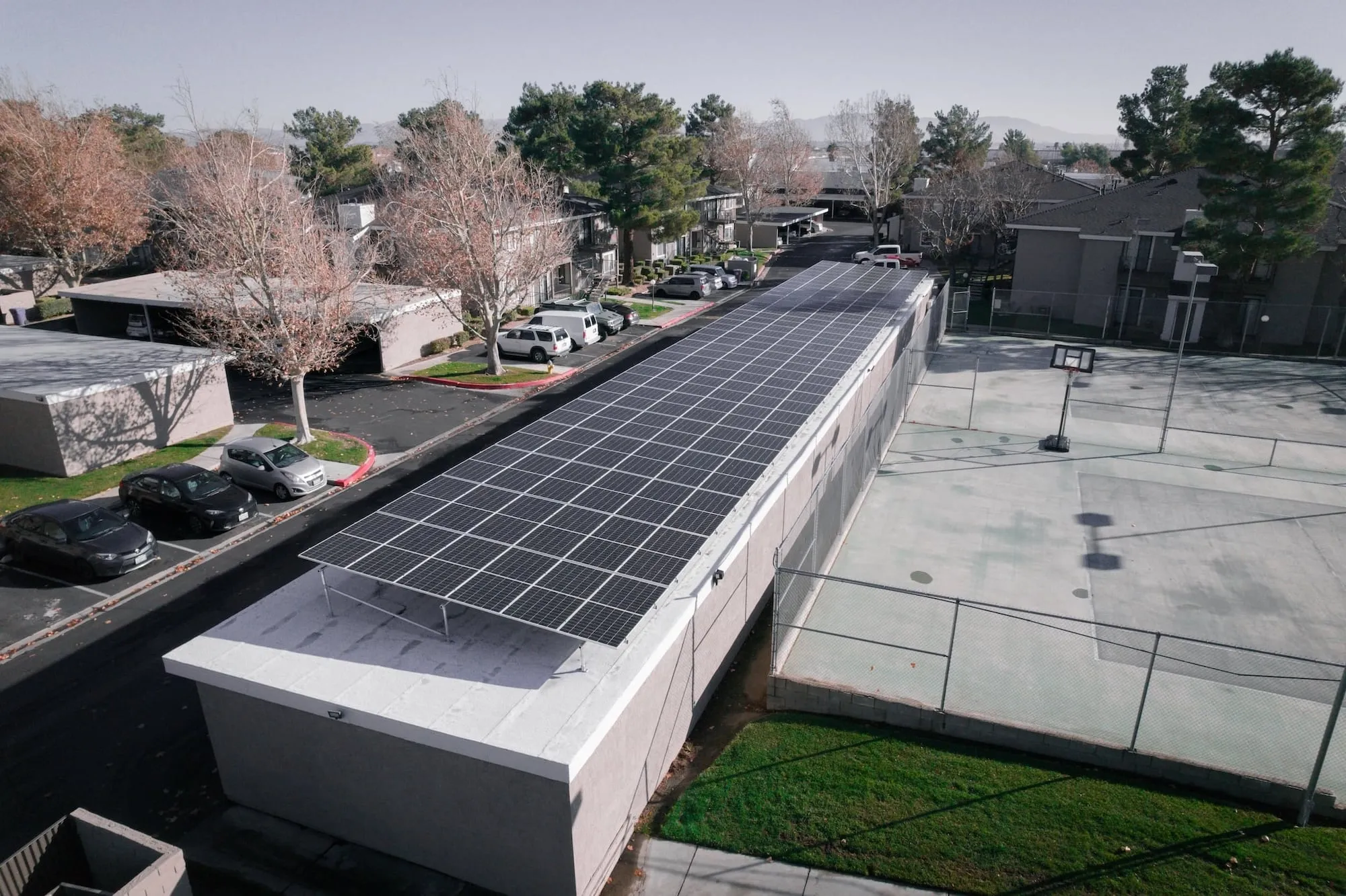 A long solar array on the flat roof of a low building beside a parking lot and tennis courts