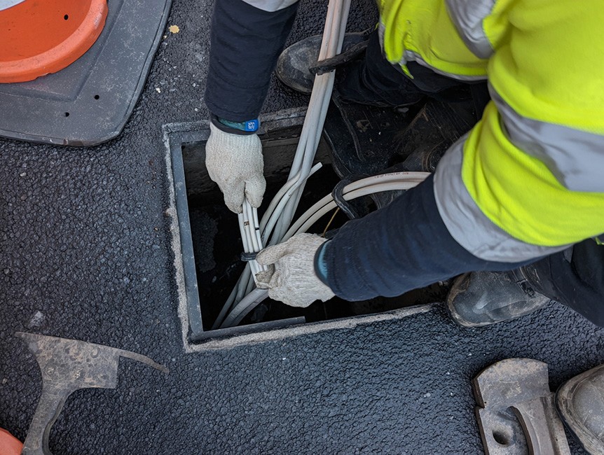 A worker in a high-visibility jacket and gloves is installing electrical cables in an underground utility access on a paved surface, surrounded by tools and safety equipment.