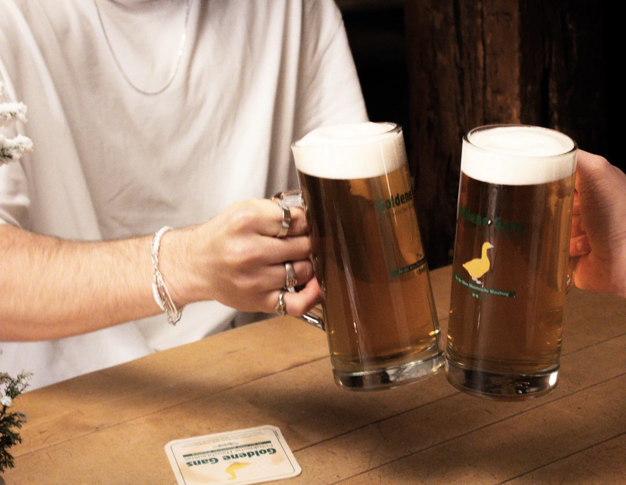 Zwei Personen stoßen mit Biergläsern an, im Hintergrund ein rustikaler Tisch.