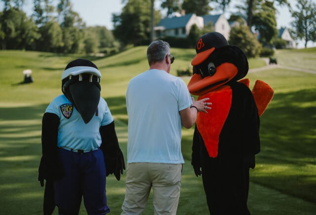 A man in a white shirt stands on a golf course talking to two bird mascots, one dressed as a police officer and the other in an orange and black outfit, during an event hosted by an advertising agency Baltimore.