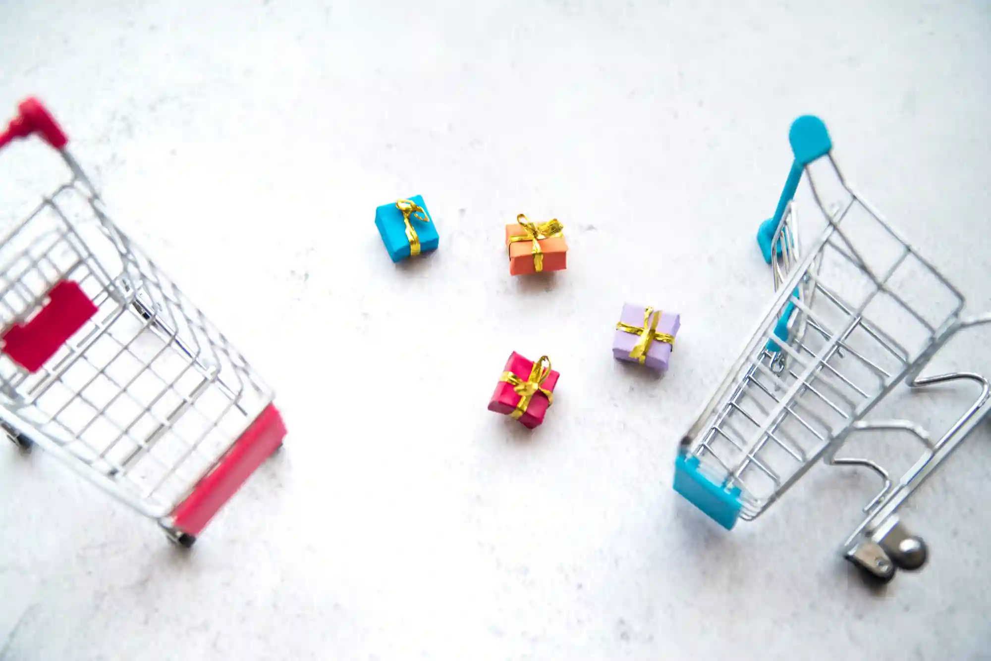 Two miniature shopping carts with tiny wrapped gift boxes in between them on a white surface.