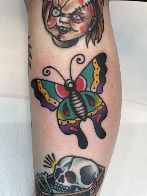Färgglad traditional-tatuering med fjäril, Chucky och skalle på underben