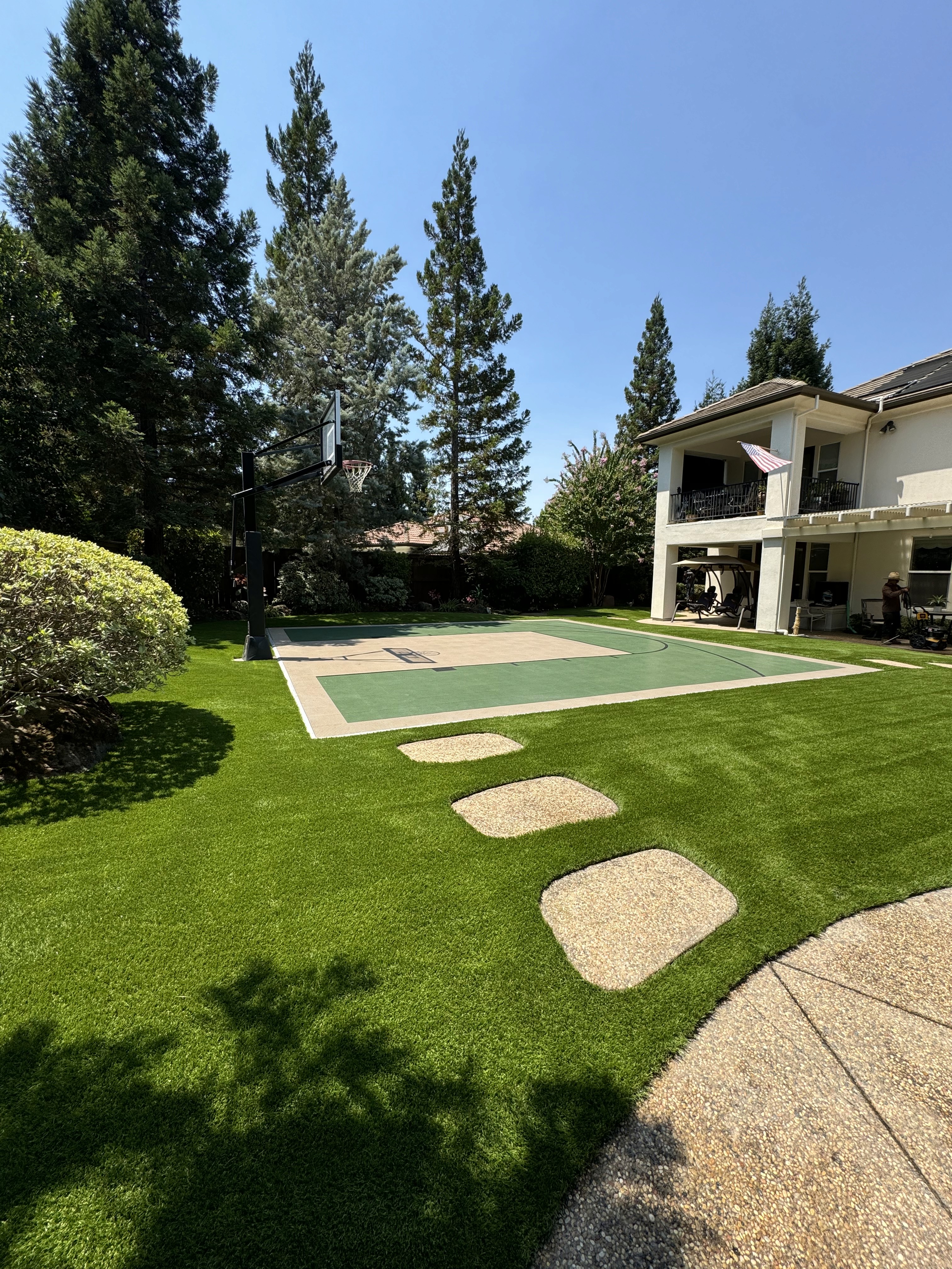 Artificial turf lawn with flagstone stepping stones and a sport court beside a stucco home framed by tall pines.