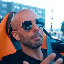 A woman with blonde bob hair in a grey coat and beige turtleneck stands against a neutral backdrop. She has a serious expression, conveying elegance.