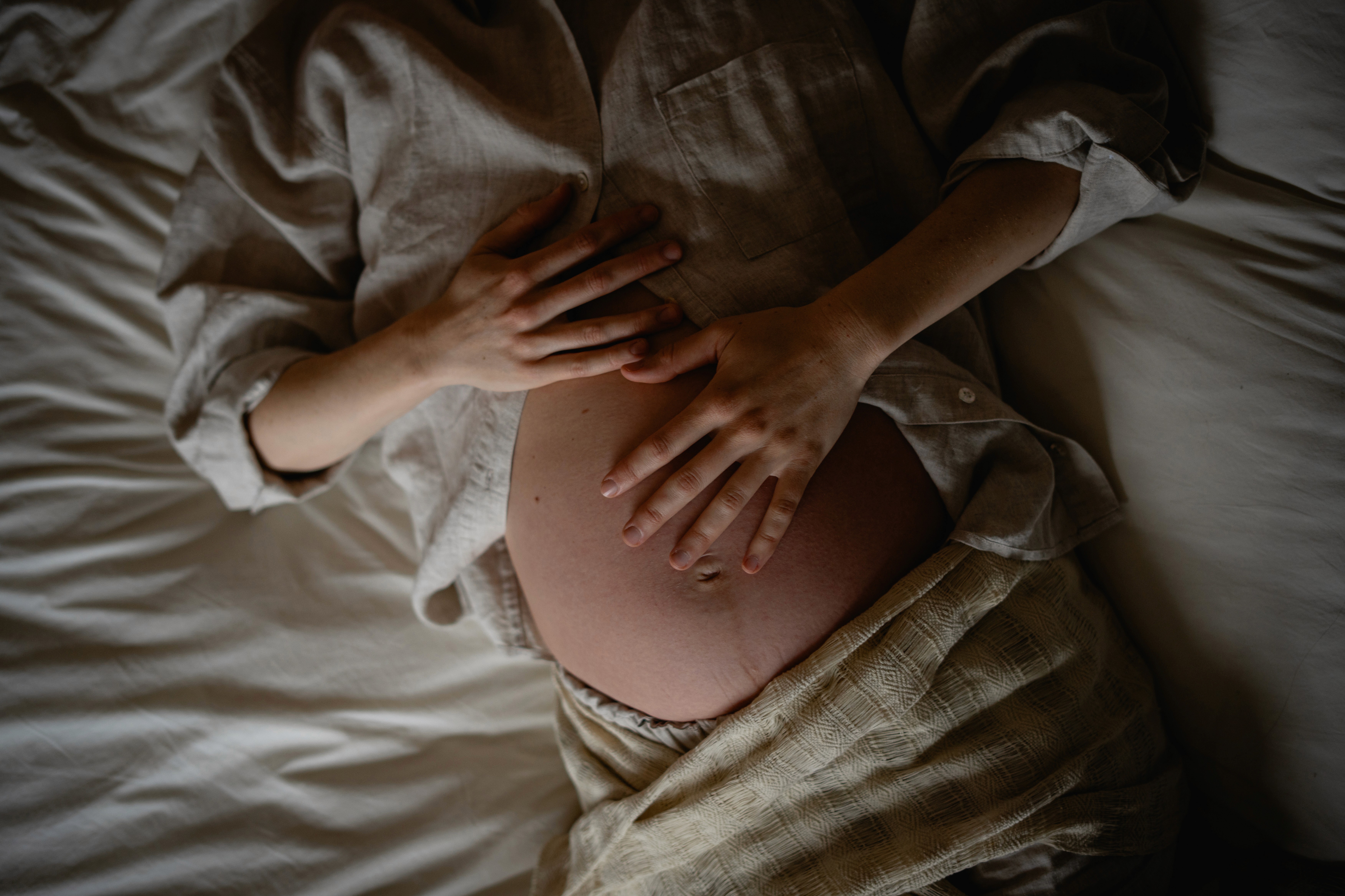 Pregnant women in linen clothes laying down on a bed with her hands on her belly