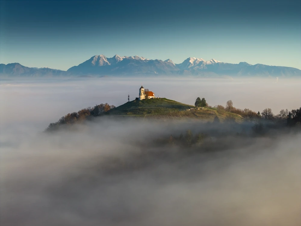 The iconic hilltop church of Sveta Ana in Slovenia, appearing as an island above a dense layer of low-lying fog during an autumn sunrise.