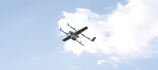 A drone with a sleek, modern design flies against a backdrop of a clear blue sky with scattered white clouds, demonstrating advanced aerial technology.
