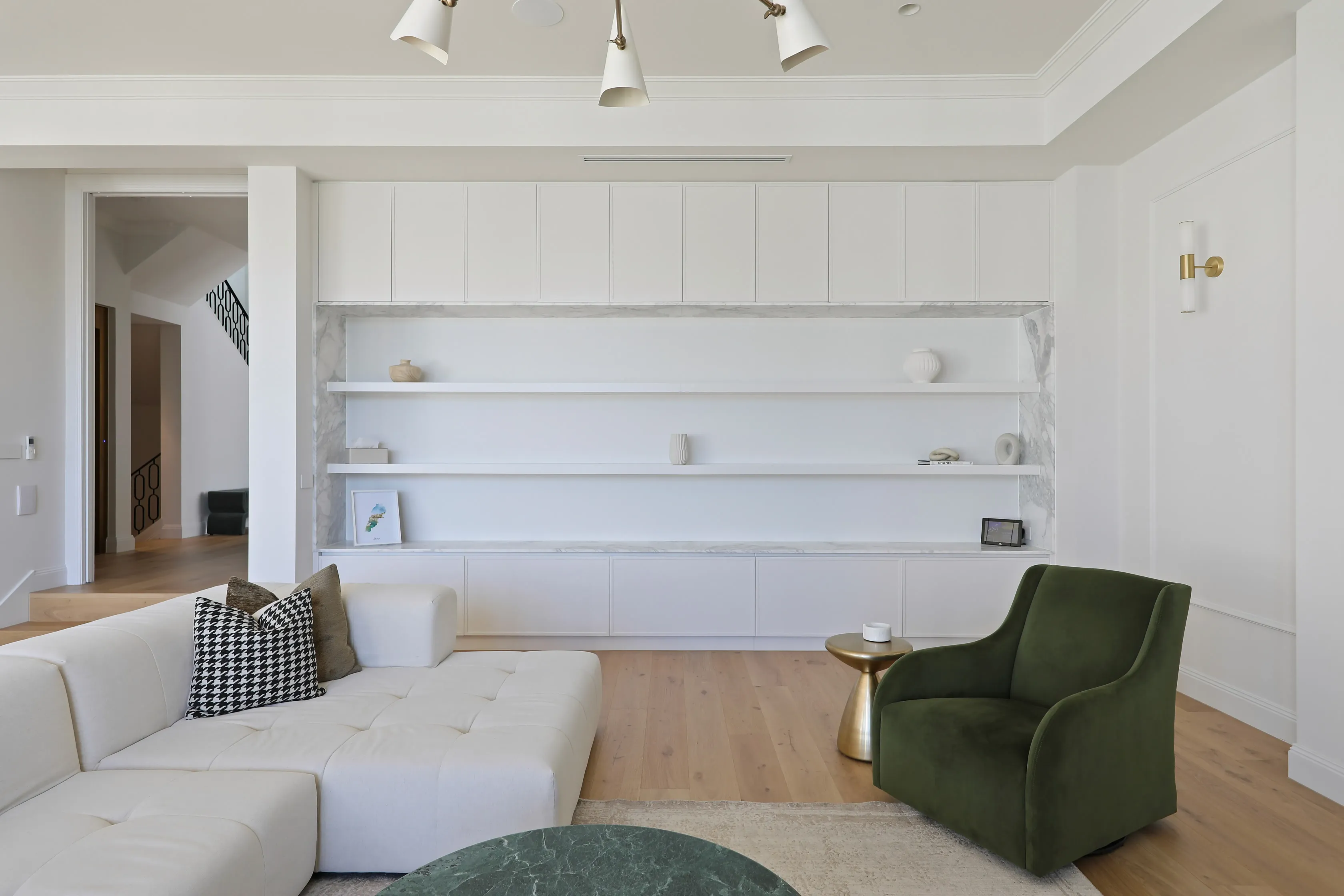 Living room with a built-in white media wall and marble-backed open shelving, a white modular sofa with houndstooth cushion, olive velvet armchair and a view through to the staircase